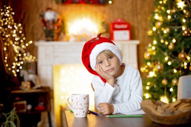 Güzel sarışın çocuk, genç okul çocuğu süslü bir evde Noel Baba 'ya mektup yazıyor süt içiyor ve kurabiye yiyor. Işıklar ve örgü oyuncaklar