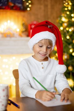 Güzel sarışın çocuk, genç okul çocuğu süslü bir evde Noel Baba 'ya mektup yazıyor süt içiyor ve kurabiye yiyor. Işıklar ve örgü oyuncaklar