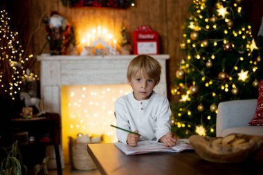 Güzel sarışın çocuk, genç okul çocuğu süslü bir evde Noel Baba 'ya mektup yazıyor süt içiyor ve kurabiye yiyor. Işıklar ve örgü oyuncaklar
