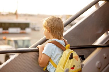 Sevimli sarışın çocuk, sırt çantalı çocuk, gün batımında havaalanında uçağa binmiş, dışarıdan manzaranın tadını çıkarıyor.