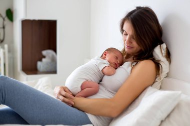 Genç anne yeni doğmuş bebeğiyle yatakta yatıyor, onu kollarına alıyor ve mutluluktan gülümsüyor.