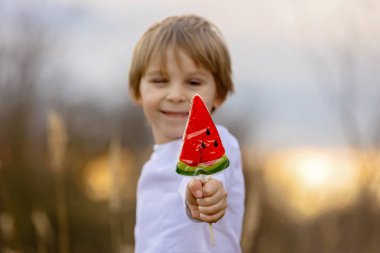 Tatlı çocuk, güzel sarışın çocuk, gün batımında parkta karpuz lolipop yiyor, açık havada güzel bir bahar havası var.