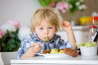 Küçük çocuk, sarışın çocuk, evde haşlanmış sebze, brokoli, patates ve havuç yerken tabletten film izliyor.