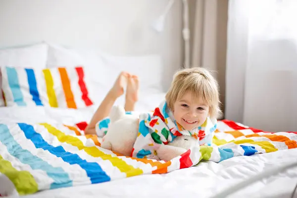 幸せな幼児の子供 カラフルなバスローブを着たブロンドの男の子 バスの後にぬいぐるみでベッドに座って カラフルな寝具 — ストック写真