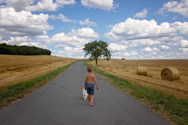 Güzel sarışın çocuk, evlat, kırsal yolda tatlı küçük köpeğiyle yürüyor. Şaşırtıcı manzara, bulutlu kırsal manzara, ağaç ve boş yol yaz zamanı, yolun yanındaki saman yığını, Portekiz