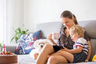 Anne ve çocuk, küçük çocuk, tablette film izliyor, masada pasta ve çiçekler.