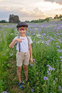 Güzel bir okul çocuğu, gün batımında çiçek tarlasında yüzü boyalı bir çocuk, elinde çiçekler ve eski moda valizlerle, yaz zamanı.