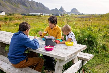 Çocuklar, Norveç, Lofoten ve Vaeroy 'da kamp yaparken ahşap bir bankta öğle yemeği yiyorlar.