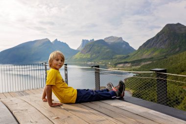 Çocuklar, 44 metre uzunluğundaki izleme platformunun kenarında durarak, Bergsfjord 'un suyuna bakarak, Kuzey Norveç' teki Senja adasının manzarasının tadını çıkarıyorlar.