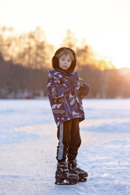 Mutlu çocuk, çocuk, sıradan giyinmiş, gün boyunca donmuş bir gölde paten kayıyor, ormanın arkasında güzel bir günbatımı, eğleniyor.