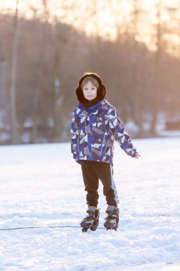 Mutlu çocuk, çocuk, sıradan giyinmiş, gün boyunca donmuş bir gölde paten kayıyor, ormanın arkasında güzel bir günbatımı, eğleniyor.