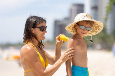 Anne ve oğlu, tatlı sarışın çocuk, denizde oynuyor, şapka, güneş gözlüğü, mayo, güneş koruyucu krem, deniz ve kum arka planı. Kavram: Aşk, yaşam tarzı, çocuklar, tatiller, çocuklar