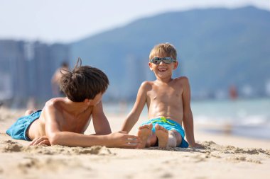 Çocuk, kumsalda ayaklarında tüyle gıdıklanan kardeş, çocuk kumla kaplı, gülümseyen, gülen, eğlenen