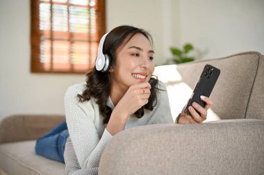 Mutlu ve güzel Asyalı bir kadın kulaklık takıyor, akıllı telefonundan müzik listesi seçiyor, en az oturma odasında kanepede yatarken müzik dinliyor..