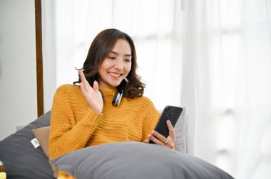Mutlu ve neşeli, rahat süveter giyen Asyalı bir kadın yataktadır, akıllı telefonuyla arkadaşlarıyla bir video görüşmesi yapıyordur..