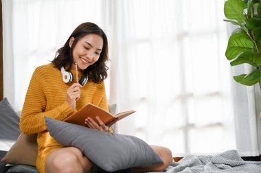 Güzel, çekici, genç Asyalı kadın, günlük giysiler içinde oturma odasında dinlenirken kitap okuyor..