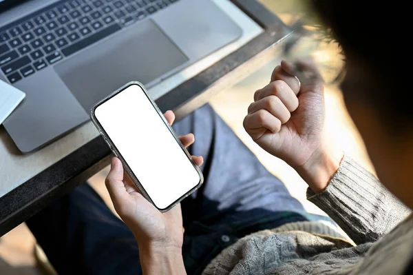 一位身穿休闲装的亚洲年轻人坐在办公桌前 用智能手机 握紧拳头 好消息 电话白屏模拟 顶视图 — 图库照片