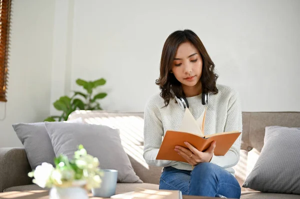专心致志的年轻亚洲女人坐在客厅的沙发上看书或研究课本信息 — 图库照片