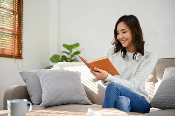 迷人的千年亚洲女人一边看书 一边在狭小的客厅里放松地坐在沙发上 知识和业余爱好概念 — 图库照片