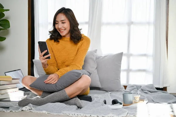 stock image Charming young Asian woman in casual clothes and comfy socks using her smartphone, looking at screen, scrolling on phone while relaxing in her living room.