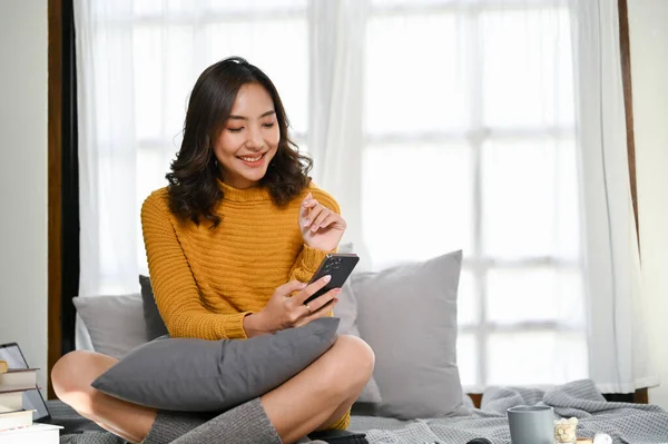 stock image Beautiful young Asian female in comfy sweater using her smartphone while sitting in the living room. chatting, online shopping, using app, reading online.