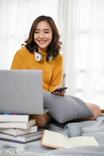 Attractive and cheerful young Asian woman remote working from home, using laptop computer, working on her tasks on bed in her minimal bedroom.