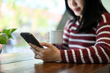 Akıllı telefonunu kullanan, internetten haber okuyan, sosyal medyasını kontrol eden ve kahvesini yudumlayan çekici bir Asyalı kadın. kırpılmış resim