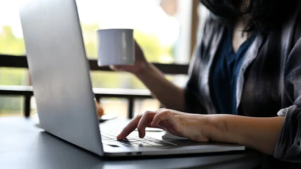 An Asian female freelancer remote working at the coffee shop, sipping morning coffee while working on her tasks on laptop computer. side view, close-up image