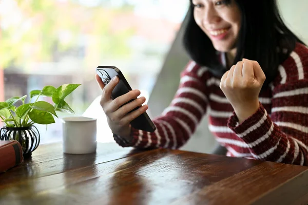 兴奋而快乐的年轻亚洲女人听到一个意想不到的消息 看着她的智能手机屏幕 坐在咖啡店里举手表决 — 图库照片