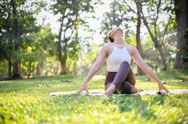 Çekici Asyalı kadın spor kıyafetleriyle Lotus pozu veya diz boyu poz verip boynunu uzatıyor, güzel yeşil parkta yoga eğitimi veriyor..