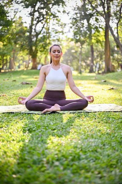 Portrait, Attractive and healthy Asian woman in sportswear meditating, concentrating on her breath, doing Lotus pose, practicing yoga in the park.