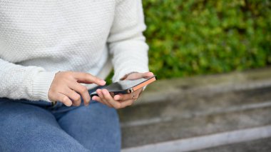 Parkta otururken akıllı telefon kullanan genç bir kadının görüntüsünü kapat. İnsanlar ve teknoloji kavramı