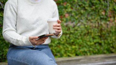 Elinde bir fincan kahveyle parkta bir bankta oturan bir kadının elinde akıllı telefonu yakın çekim görüntüsü..