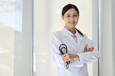 Profesyonel ve güzel Asyalı kadın doktor, kliniğinde duruyor, steteskop tutuyor, kollarını kavuşturuyor, gülümsüyor ve kameraya bakıyor..