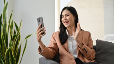 Kanepede oturan, el sallayan, akıllı telefonuna rağmen bir video görüşmesinde konuşan yakışıklı Asyalı genç iş kadını..