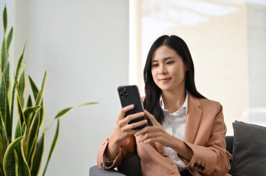 Resmi takım elbiseli güzel Asyalı iş kadını ya da bayan patron kanepede oturuyor ve akıllı telefonunu kullanıyor..