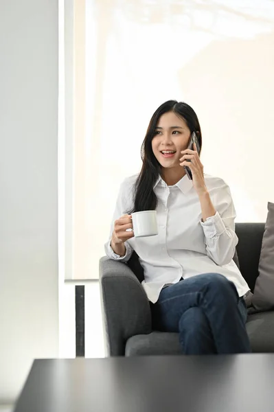 有魅力的年轻亚洲女人在沙发上喝咖啡时 一边和朋友通电话 一边表现自己的风采 — 图库照片