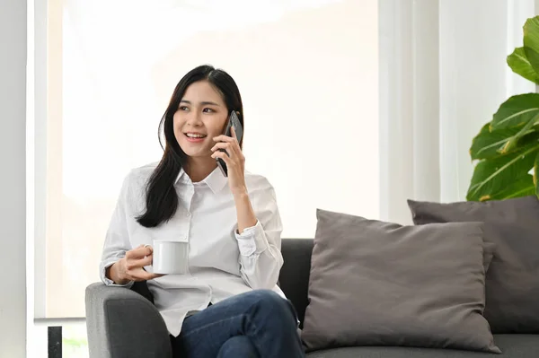 迷人的年轻亚洲女人一边和朋友通电话 一边在舒适的沙发上喝咖啡 — 图库照片