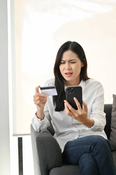 不快乐和不满意的亚洲女人坐在沙发上 看着她的智能手机屏幕 对移动银行的应用有问题 具有电话和信用卡概念的女性 — 图库照片