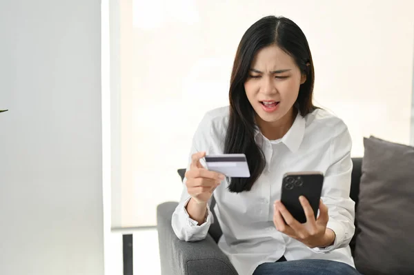 不满意的亚洲女人坐在沙发上 看着她的智能手机屏幕 对移动银行的应用有问题 网上支付 网上购物 — 图库照片