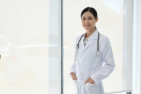 制服を着たプロの若いアジアの女性医師 病院に立って ポケットに手 笑顔とカメラを見て — ストック写真