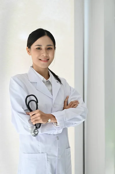 身穿白衣的亚洲职业女性医生 站在诊所里 手持听诊器 双臂交叉 面带微笑地看着镜头 — 图库照片