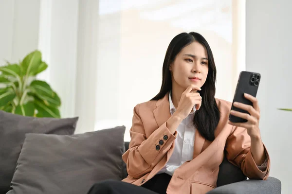 漂亮的年轻亚洲女商人或女经理坐在沙发上 看着她的智能手机屏幕 沉思或计划着什么 — 图库照片