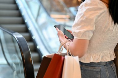 Kesilmiş fotoğraf, telefonuyla ve alışveriş çantasıyla bir sonraki kata çıkan Asyalı genç bayan alışveriş merkezinden alışveriş yapıyor..