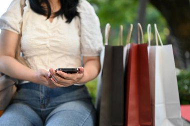 Güzel, Asyalı bir kadın günlük kıyafetleriyle akıllı telefon kullanıyor alışveriş merkezindeki bir bankta alışveriş torbalarıyla oturuyor. kırpılmış resim