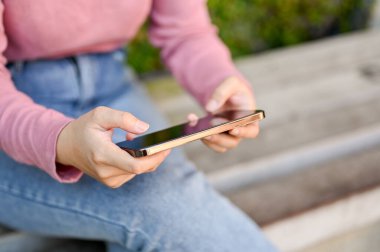 Rahat genç Asyalı kadın akıllı telefonunu video izlemek ya da parkta bankta otururken mobil oyun oynamak için kullanıyor. kırpılmış ve yakın plan resim