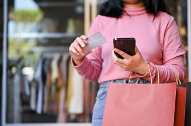 Keskin nişancı, elinde kredi kartı ve akıllı telefonu olan genç Asyalı bir kadın elbise mağazasının önünde dikiliyor..