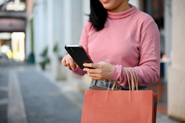 Genç Asyalı kadın alışveriş çantalarını taşıyor ve şehir meydanında alışveriş yaparken akıllı telefonunu kullanıyor. kırpılmış atış