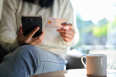 Kesilmiş atış, kafeteryada otururken akıllı cep telefonu ve kredi kartını tutan beyaz kazaklı genç Asyalı kadın. çevrimiçi alışveriş kavramı