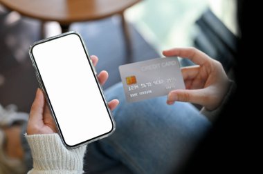 Kafede oturan Asyalı bir kadın, elinde akıllı telefonu ve kredi kartıyla. telefon beyaz ekran modeli.